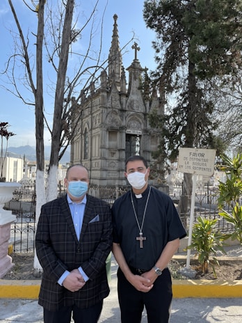 Two men wearing face masks stand in front of an ornate, Gothic-style mausoleum surrounded by trees. One man is wearing a plaid suit jacket and the other is dressed in clergy attire with a cross necklace. A sign in Spanish is visible on the right side.