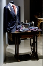 A tailored suit with a red patterned tie is displayed on a mannequin. An old-fashioned sewing machine sits beside it, along with several folded fabrics, suggesting a tailor's workspace. The setting has a classic and elegant atmosphere.