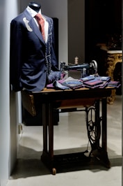 A tailored suit with a red patterned tie is displayed on a mannequin. An old-fashioned sewing machine sits beside it, along with several folded fabrics, suggesting a tailor's workspace. The setting has a classic and elegant atmosphere.
