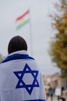 A person is covered with an Israeli flag, facing away from the camera. In the background, there is a blurred flag with red, white, and green horizontal stripes with a yellow symbol in the center. The scene appears to be outdoors, with some trees and a cloudy sky.