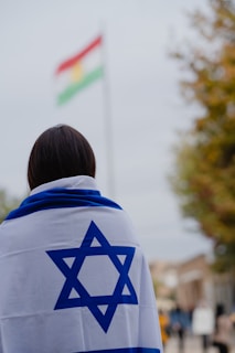A person is covered with an Israeli flag, facing away from the camera. In the background, there is a blurred flag with red, white, and green horizontal stripes with a yellow symbol in the center. The scene appears to be outdoors, with some trees and a cloudy sky.
