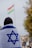 A person is covered with an Israeli flag, facing away from the camera. In the background, there is a blurred flag with red, white, and green horizontal stripes with a yellow symbol in the center. The scene appears to be outdoors, with some trees and a cloudy sky.