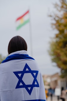 A person is covered with an Israeli flag, facing away from the camera. In the background, there is a blurred flag with red, white, and green horizontal stripes with a yellow symbol in the center. The scene appears to be outdoors, with some trees and a cloudy sky.