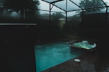 A dimly lit indoor swimming pool area features a pool and adjacent hot tub with light ripples and bubbles on the surface. The space is enclosed with dark glass creating a moody atmosphere, and outside, trees and a cloudy sky are faintly visible.