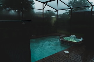 A dimly lit indoor swimming pool area features a pool and adjacent hot tub with light ripples and bubbles on the surface. The space is enclosed with dark glass creating a moody atmosphere, and outside, trees and a cloudy sky are faintly visible.