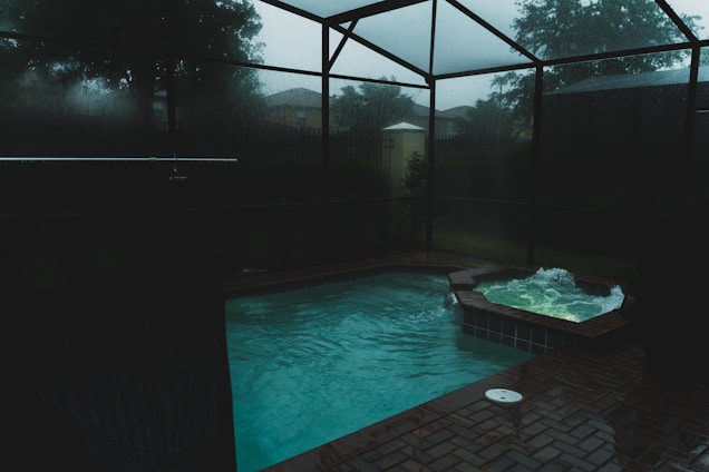A dimly lit indoor swimming pool area features a pool and adjacent hot tub with light ripples and bubbles on the surface. The space is enclosed with dark glass creating a moody atmosphere, and outside, trees and a cloudy sky are faintly visible.