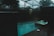 A dimly lit indoor swimming pool area features a pool and adjacent hot tub with light ripples and bubbles on the surface. The space is enclosed with dark glass creating a moody atmosphere, and outside, trees and a cloudy sky are faintly visible.