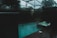 A dimly lit indoor swimming pool area features a pool and adjacent hot tub with light ripples and bubbles on the surface. The space is enclosed with dark glass creating a moody atmosphere, and outside, trees and a cloudy sky are faintly visible.