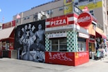 A vibrant street scene featuring a pizza stand named Gabriella's, with a bold red and white color scheme. The adjacent mural depicts several people with the phrase 'Born x Raised' in large letters. Nearby are shops with colorful storefronts.