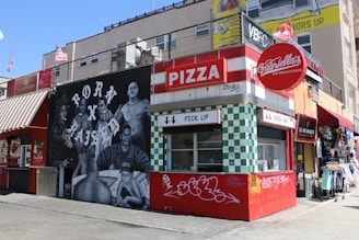 A vibrant street scene featuring a pizza stand named Gabriella's, with a bold red and white color scheme. The adjacent mural depicts several people with the phrase 'Born x Raised' in large letters. Nearby are shops with colorful storefronts.