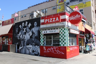 A vibrant street scene featuring a pizza stand named Gabriella's, with a bold red and white color scheme. The adjacent mural depicts several people with the phrase 'Born x Raised' in large letters. Nearby are shops with colorful storefronts.