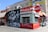 A vibrant street scene featuring a pizza stand named Gabriella's, with a bold red and white color scheme. The adjacent mural depicts several people with the phrase 'Born x Raised' in large letters. Nearby are shops with colorful storefronts.