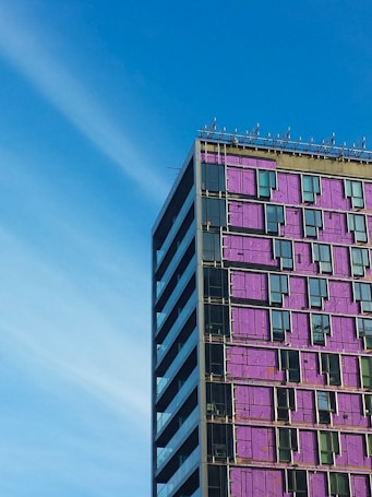 A tall, rectangular building with a modern design is partly covered in purple cladding. The structure features multiple rows of windows and what appears to be construction work in progress. The sky is clear and blue, suggesting a bright day.