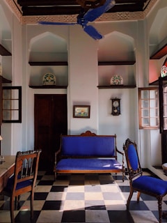 An elegant, vintage-style room with high ceilings and decorative arches. The room features a checkered black and white tile floor, a wooden sofa, and chairs with vibrant blue upholstery. Intricate carvings are visible on the ceiling, and there is a blue ceiling fan. A grandfather clock and framed artwork adorn the walls, while ornate pottery sits on recessed wall shelves.
