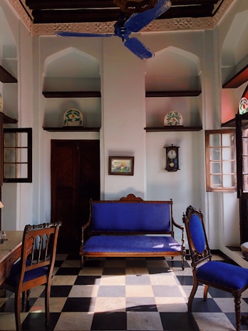 An elegant, vintage-style room with high ceilings and decorative arches. The room features a checkered black and white tile floor, a wooden sofa, and chairs with vibrant blue upholstery. Intricate carvings are visible on the ceiling, and there is a blue ceiling fan. A grandfather clock and framed artwork adorn the walls, while ornate pottery sits on recessed wall shelves.