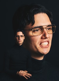 A man with dark hair and glasses appears in two positions within the frame. The larger portrait shows a close-up of his face with a slight smile, while the smaller image features him sitting with hands folded, wearing a black turtleneck against a dark background.