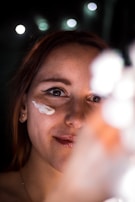 A woman smiles softly as she applies cream to her face. Her skin appears smooth and her lips are slightly glossy. The background is blurred, with bokeh lights creating a soft, ambient glow.