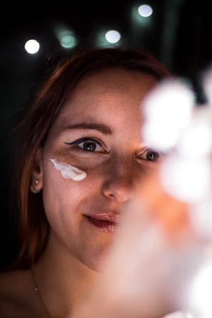 A woman smiles softly as she applies cream to her face. Her skin appears smooth and her lips are slightly glossy. The background is blurred, with bokeh lights creating a soft, ambient glow.