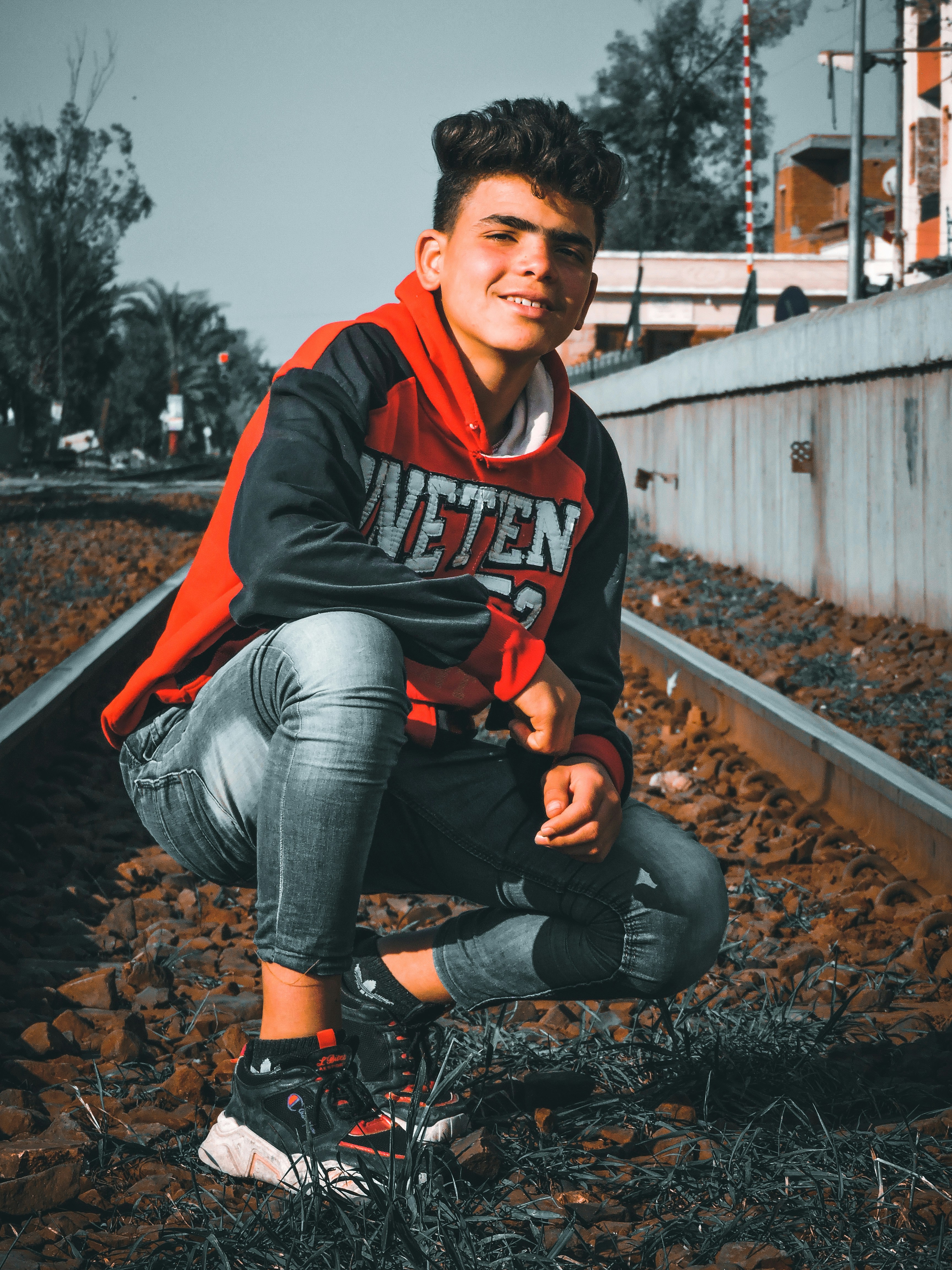 Pria dengan hoodie merah dan hitam dan jeans denim abu-abu duduk di rel kereta pada siang hari