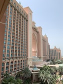 A grand hotel building with a large central archway and adjacent tall towers. The architecture has a Middle Eastern influence with ornate detailing and decorative elements. The facade is a sandy pink color, complementing the lush greenery of palm trees and landscaped gardens at the base of the structure.