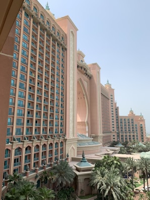 A grand hotel building with a large central archway and adjacent tall towers. The architecture has a Middle Eastern influence with ornate detailing and decorative elements. The facade is a sandy pink color, complementing the lush greenery of palm trees and landscaped gardens at the base of the structure.