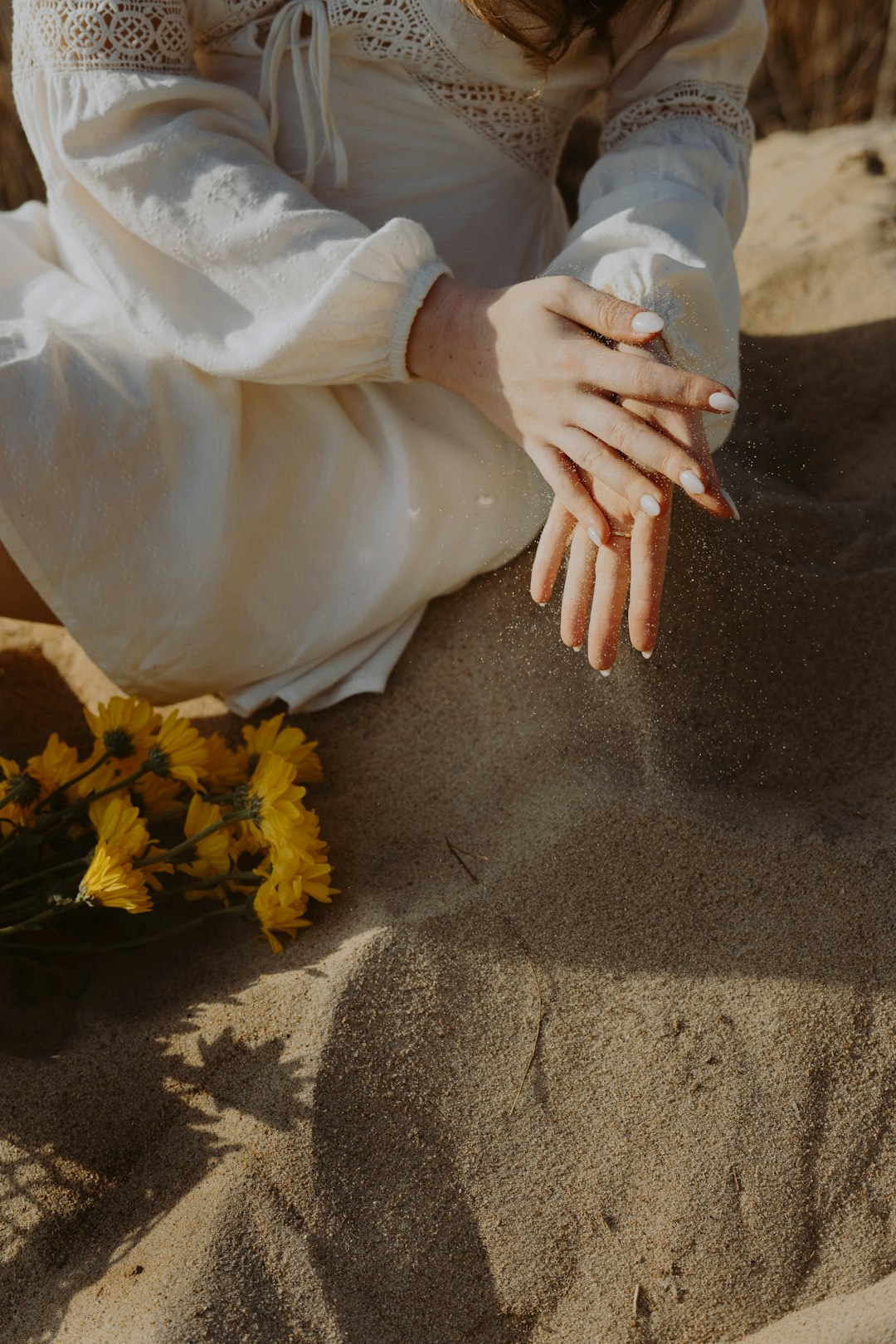 person in white long sleeve shirt and white pants sitting on sand with yellow flowers