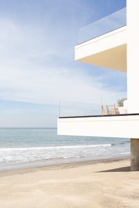A modern building with glass railings overlooks a sandy beach and calm ocean. The architecture is minimalist with a focus on clean lines and an open view.