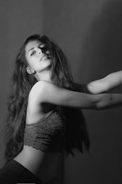 A black and white photograph of a woman with long hair in a detailed lace top, striking a pose with an artistic flair. The lighting creates dramatic shadows, adding depth and highlighting her features.
