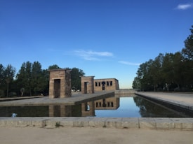 An ancient stone temple is reflected in a large rectangular pool of water, surrounded by a park with lush green trees against a clear blue sky.