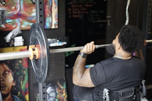 A person in a gym setting is lifting a barbell with weight plates while seated in a wheelchair. The person's back is turned, showing muscular arms, tattoos, and an afro hairstyle. The surroundings have artistic paintings on the walls and gym equipment.
