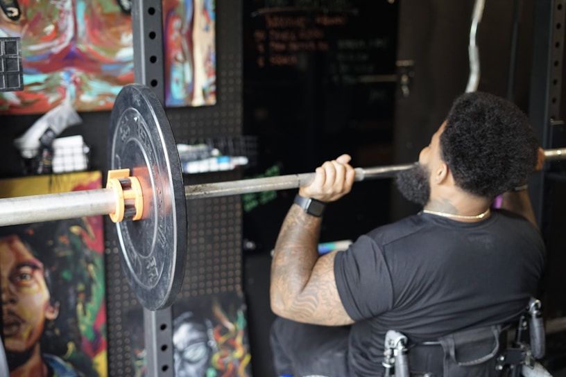 A person in a gym setting is lifting a barbell with weight plates while seated in a wheelchair. The person's back is turned, showing muscular arms, tattoos, and an afro hairstyle. The surroundings have artistic paintings on the walls and gym equipment.