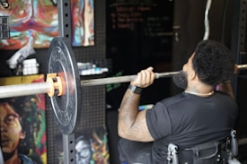 A person in a gym setting is lifting a barbell with weight plates while seated in a wheelchair. The person's back is turned, showing muscular arms, tattoos, and an afro hairstyle. The surroundings have artistic paintings on the walls and gym equipment.