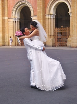 A couple in wedding attire is in an affectionate pose with the groom lifting the bride, surrounded by an elegant architectural background. The bride wears a flowing white dress and holds a bouquet of pink flowers, while the groom is dressed in a light-colored suit.