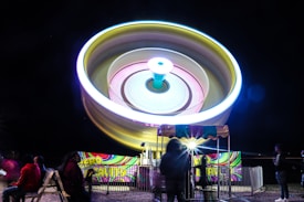 A brightly lit amusement ride spins rapidly, creating a circular blur of colors against the night sky. The ride resembles a giant spinning top, with vibrant neon lights creating concentric circles. Below, people are gathered in semi-darkness, some seated and others standing near the entrance gate.