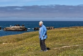An elderly person with silver hair, dressed in a blue jacket and dark pants, stands on a grassy hillside with a walking stick. Yellow wildflowers are scattered across the grass. In the background, the ocean stretches out under a cloudy sky, with rocky formations jutting out of the water.
