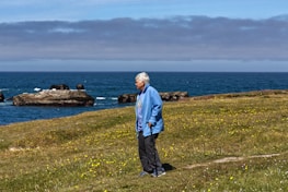 An elderly person with silver hair, dressed in a blue jacket and dark pants, stands on a grassy hillside with a walking stick. Yellow wildflowers are scattered across the grass. In the background, the ocean stretches out under a cloudy sky, with rocky formations jutting out of the water.