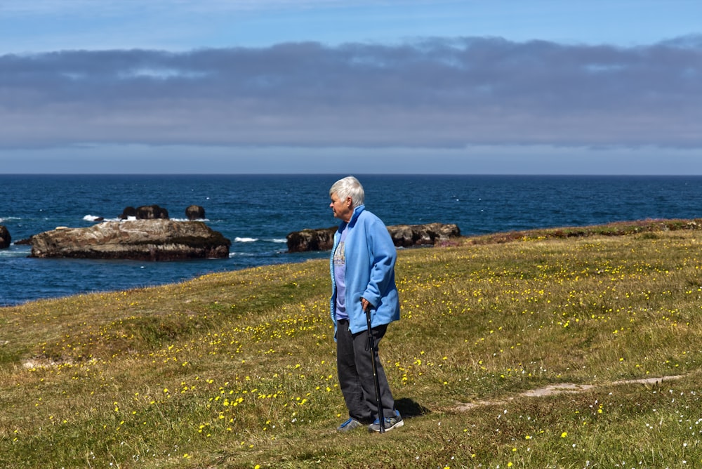 homem na jaqueta azul em pé no campo de grama verde perto do corpo de água durante o dia