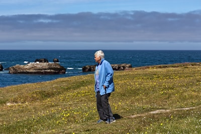 An elderly person with silver hair, dressed in a blue jacket and dark pants, stands on a grassy hillside with a walking stick. Yellow wildflowers are scattered across the grass. In the background, the ocean stretches out under a cloudy sky, with rocky formations jutting out of the water.