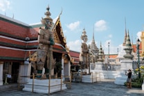 An ornate architectural scene showcasing two large guardian statues with intricate details and vibrant colors, standing in front of a temple with multi-layered roofs. The background includes several tall spires and decorative elements typical of Southeast Asian temple design. A person on the right is taking a photo, dressed in casual attire.