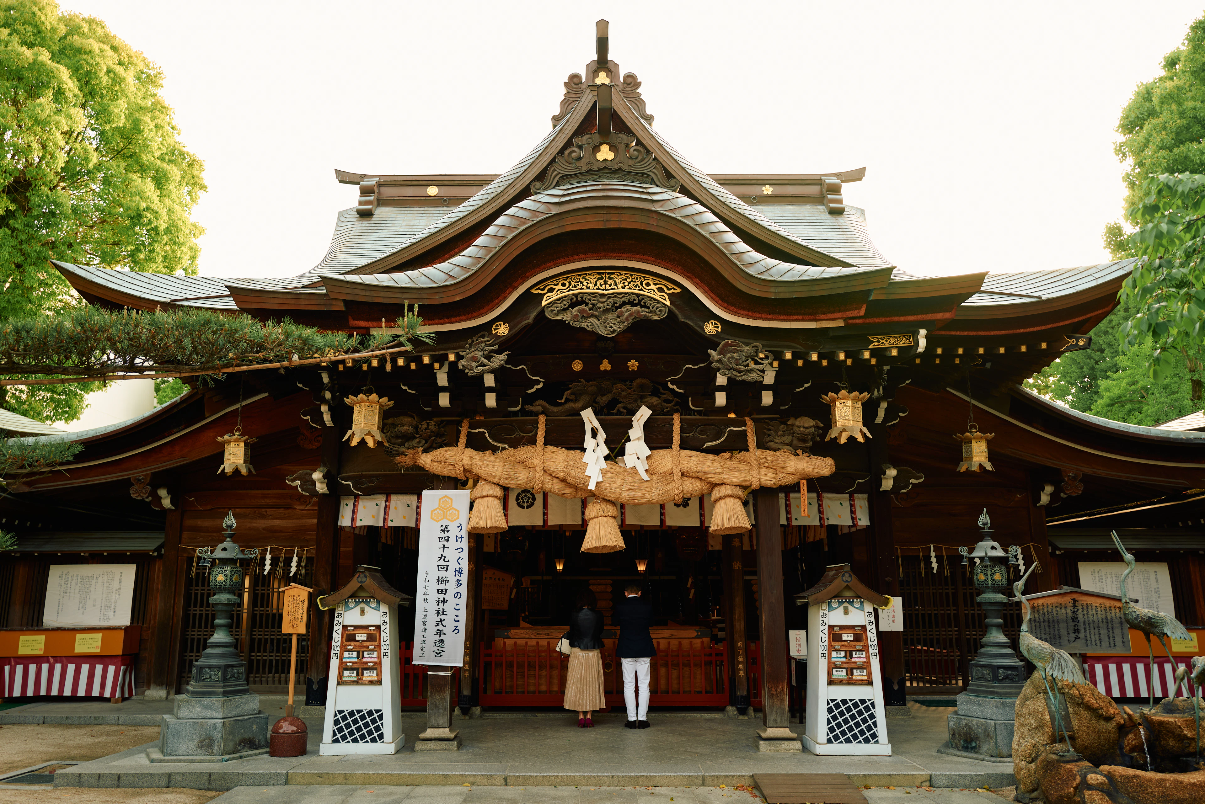 Two worshippers standing before the great braided straw rope at the main hall of Kushida Shrine in Hakata, Fukuoka, beneath its deeply carved and gilded wooden gable.