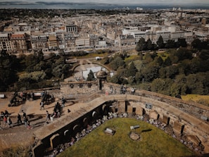 A historic cityscape with a vast array of intricately designed buildings and streets stretching into the distance. The foreground features an ancient stone structure and walkway with people casually walking, taking photos, and exploring the area. Lush green trees and manicured gardens provide a contrast to the dense urban architecture.