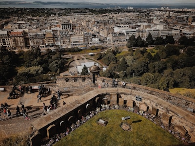 A historic cityscape with a vast array of intricately designed buildings and streets stretching into the distance. The foreground features an ancient stone structure and walkway with people casually walking, taking photos, and exploring the area. Lush green trees and manicured gardens provide a contrast to the dense urban architecture.