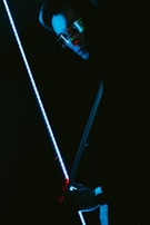 A person wearing sunglasses is holding a blue illuminated sword or stick. The lighting is dark, creating a mysterious and futuristic atmosphere with reflections of blue and red tones on the person's face and surroundings.