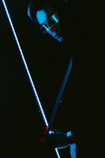 A person wearing sunglasses is holding a blue illuminated sword or stick. The lighting is dark, creating a mysterious and futuristic atmosphere with reflections of blue and red tones on the person's face and surroundings.