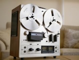 A vintage reel-to-reel tape recorder with two large reels mounted on the front. It has a metallic and wooden chassis, featuring dials, switches, and VU meters for recording and playback operations. The environment suggests an indoor setting, possibly a living room, with a soft sofa and a neutral color palette.