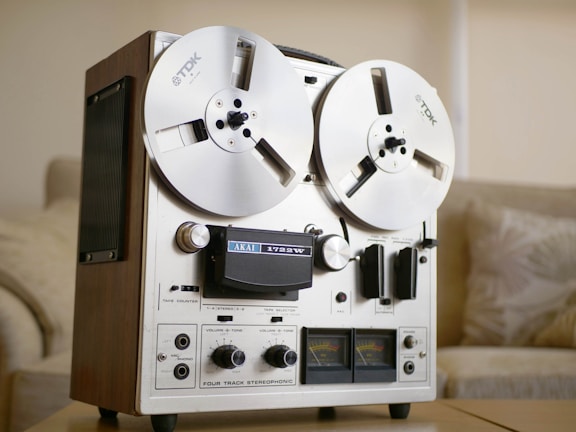 A vintage reel-to-reel tape recorder with two large reels mounted on the front. It has a metallic and wooden chassis, featuring dials, switches, and VU meters for recording and playback operations. The environment suggests an indoor setting, possibly a living room, with a soft sofa and a neutral color palette.
