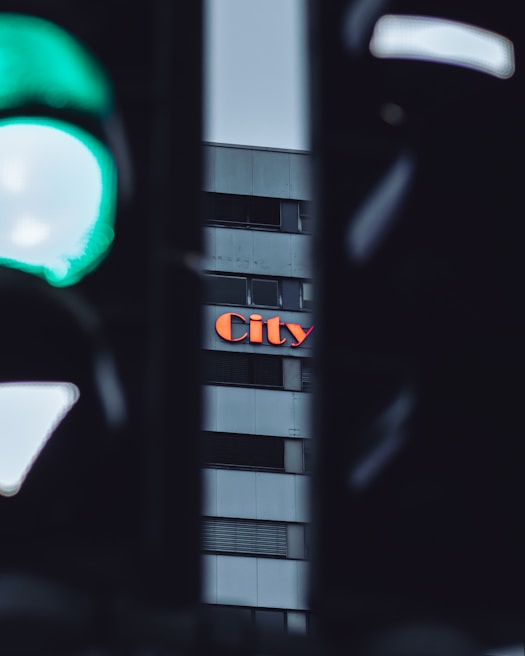 A close-up view of a building featuring the word 'City' in bright orange letters. The building's facade is segmented with horizontal lines, and a green traffic light is prominently visible in the foreground, partially blurred.