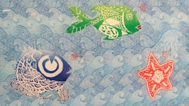 The image depicts a colorful and artistic underwater scene. There are two stylized fish and one starfish against a background of repeating blue waves. The fish are rendered in green and large stripes of blue with intricate patterns, while the starfish is red with white patterns. The overall style is illustrative and playful.