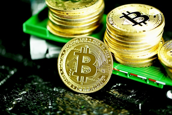 A stack of shiny gold coins with the Bitcoin logo printed prominently on each coin. The coins are placed on a green surface, reflecting light, with a prominent Bitcoin coin leaning against the stack.