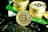 A stack of shiny gold coins with the Bitcoin logo printed prominently on each coin. The coins are placed on a green surface, reflecting light, with a prominent Bitcoin coin leaning against the stack.